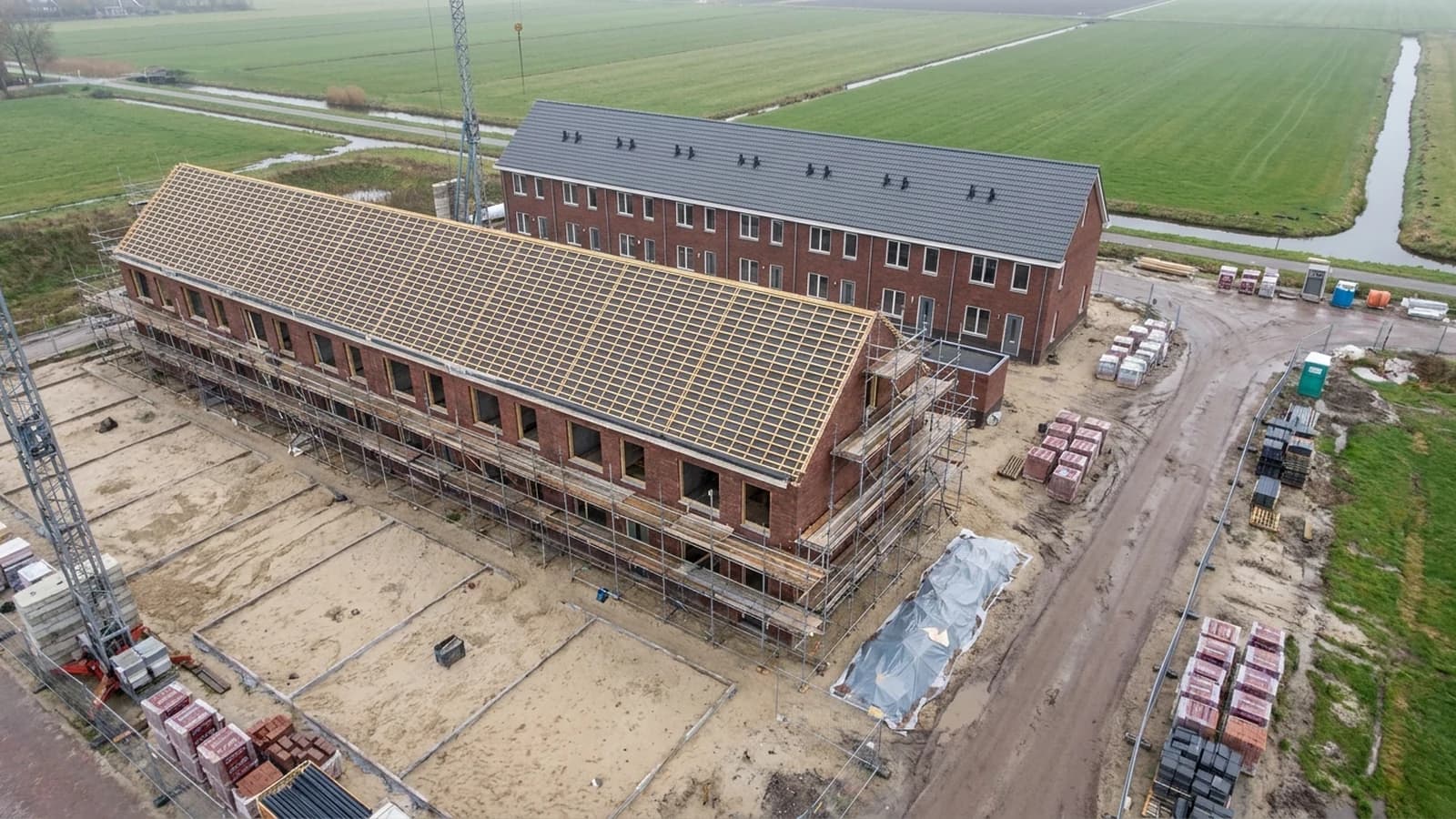 Rij nieuwbouwwoningen in aanbouw in een Nederlandse polder, met steigers en een dak in de kap